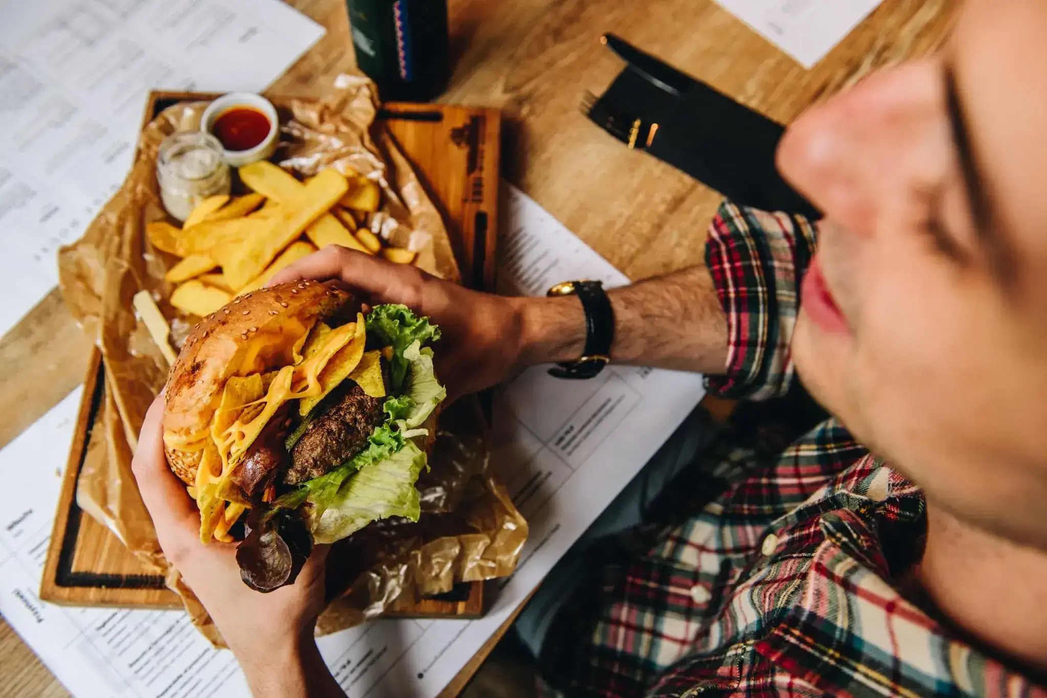 Mężczyzna trzyma w obu rękach hamburgera, którego zaraz będzie jadł. Trzyma go nad drewnianą deską, na której znajduje się papier do pieczenia, a na nim frytki belgijskie i dwa sosy – w małym słoiczku biały, a w małej białej miseczce czerwony.
