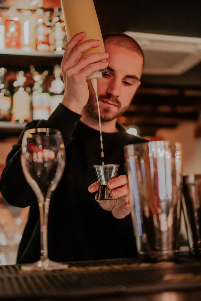 Młody barman w skupieniu odmierza składniki do drinka. Wlewa żółty płyn do metalowej miarki, przed nim stoją puste naczynia – szklanka i kieliszek.