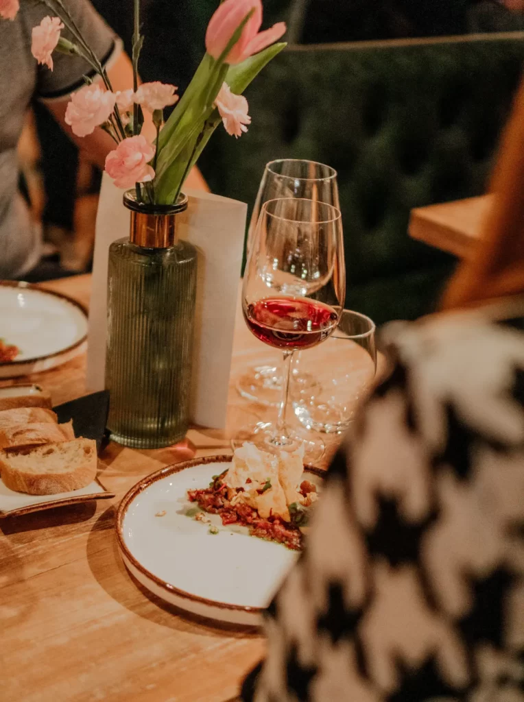 Na zdjęciu widać fragment stolika, przy którym siedzą dwie osoby. Jedna jest odwrócona tyłem. Nie widać całych postaci. Na stoliku znajdują się: dwa talerze z jedzeniem, tacka z krojonym białym chlebem, dwa kieliszki do wina. Jeden z nich jest pusty, w drugim jest czerwone wino. Centralne miejsce zajmuj mały zielony wazon, a w nim różowe kwiaty.