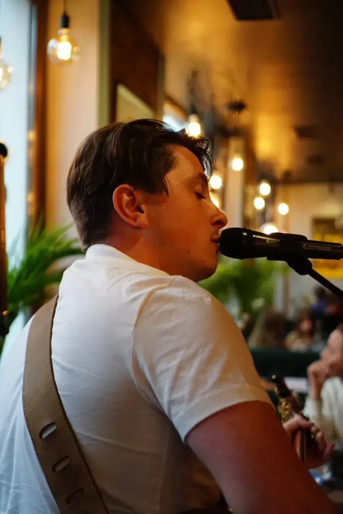 Młody muzyk w białej koszulce śpiewa do mikrofonu, tuż przy jego ustach. W tle wnętrze restauracji.