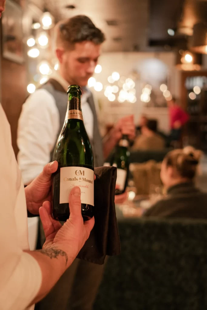 Dwie osoby przygotowują się do nalewania wina – w rękach trzymają otwarte, zielone butelki z białymi etykietami. W tle niewyraźnie sylwetki gości.