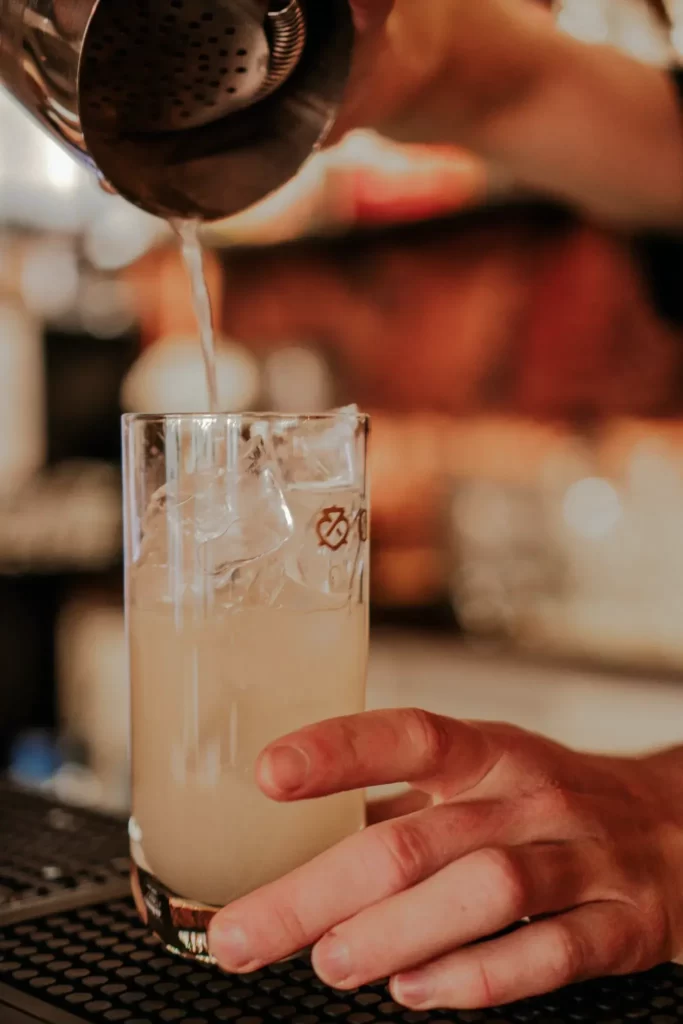 Męska dłoń trzyma wysoką szklankę, do której wlewany jest ze srebrnego szejkera jasno biały drink. Na górze drinka znajdują się kostki lodu.