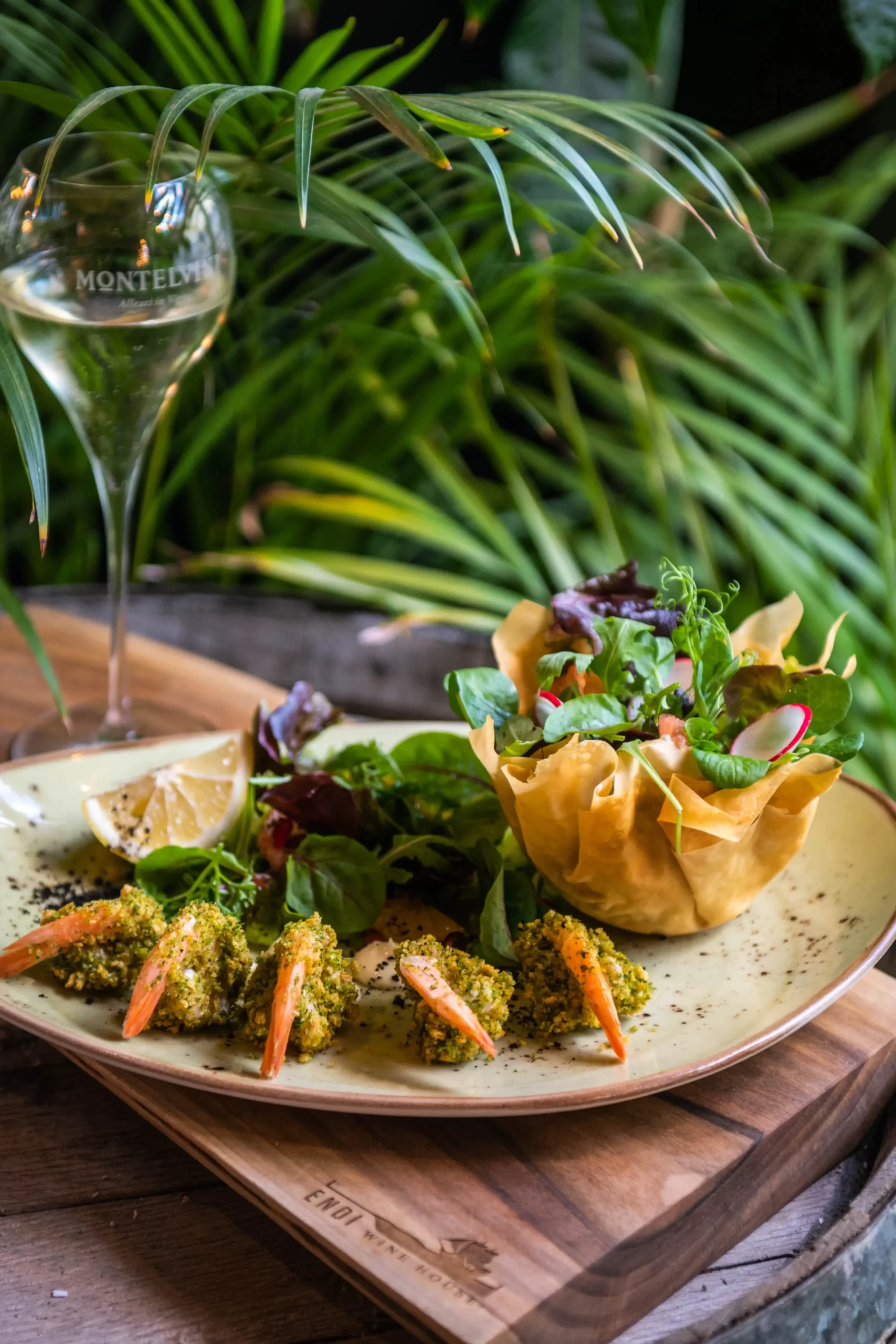 A modern plate with shrimp and a glass of white wine sits on a wine barrel, on a board with the Endi Wine House logo. In the background are green leaves of vegetation. On the plate beautifully arranged shrimp, surrounded by a piece of lemon, an edible goblet with salad.