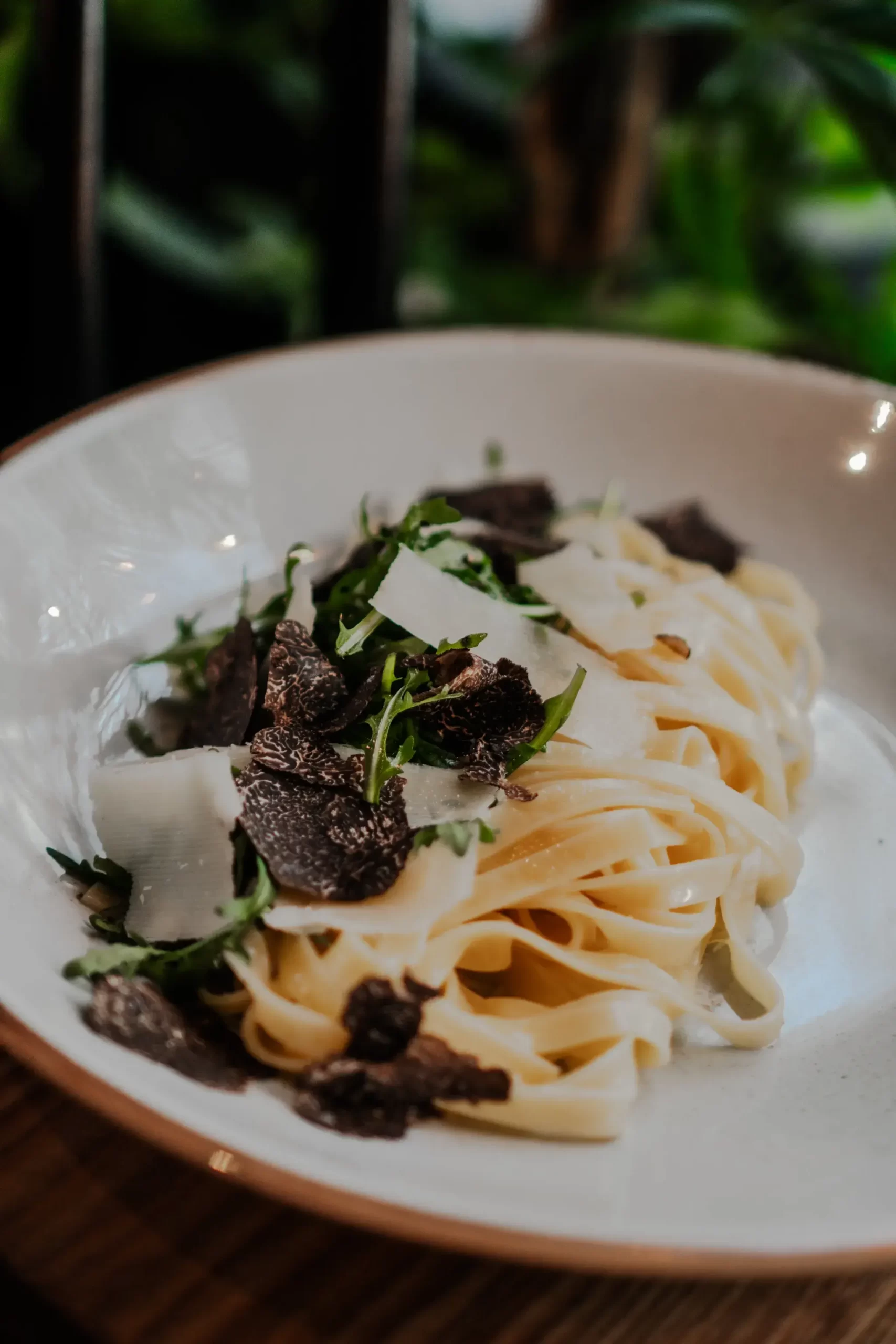 On a white plate is arranged tagliatelle with fresh truffle cut into thin slices, arugula and sliced Parmesan cheese.