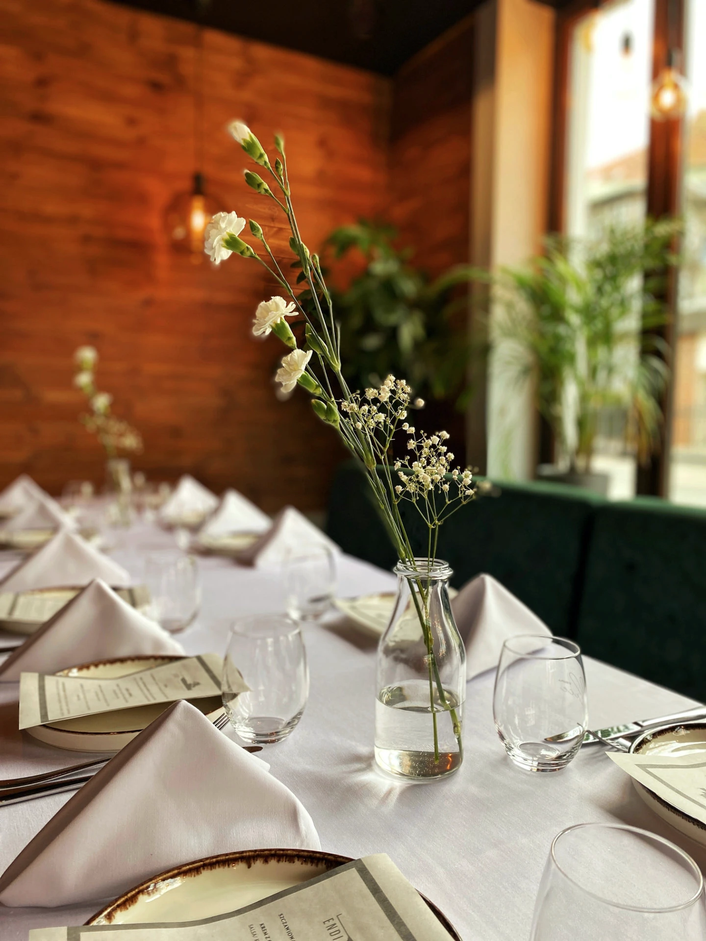 Podłużny stół w restauracji nakryty białym, klasycznym obrusem. Na stole znajdują się kremowe talerzyki, z ozdobnymi brzegami, obok nich trójkątnie złożone białe serwetki, srebrne sztućce oraz puste szklanki na napoje. Na każdym talerzyku leży menu przygotowane na uroczystość. W dwóch prostych, szklanych wazonach znajdują się delikatne, białe polne kwiaty.