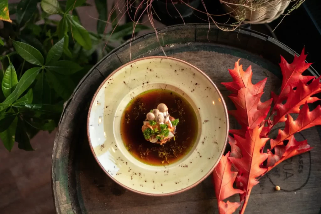Na beczce znajduje się beżowy talerz z ciemne plami z zupą brązową zupą grzybową. Na środku talerza znajduje się niesamowita dekoracja z grzybów. Talerz z zupą otoczony jest czerwonymi, jesiennymi liśćmi, po lewej stronie od beczki, znajdują się zielone rośliny.
