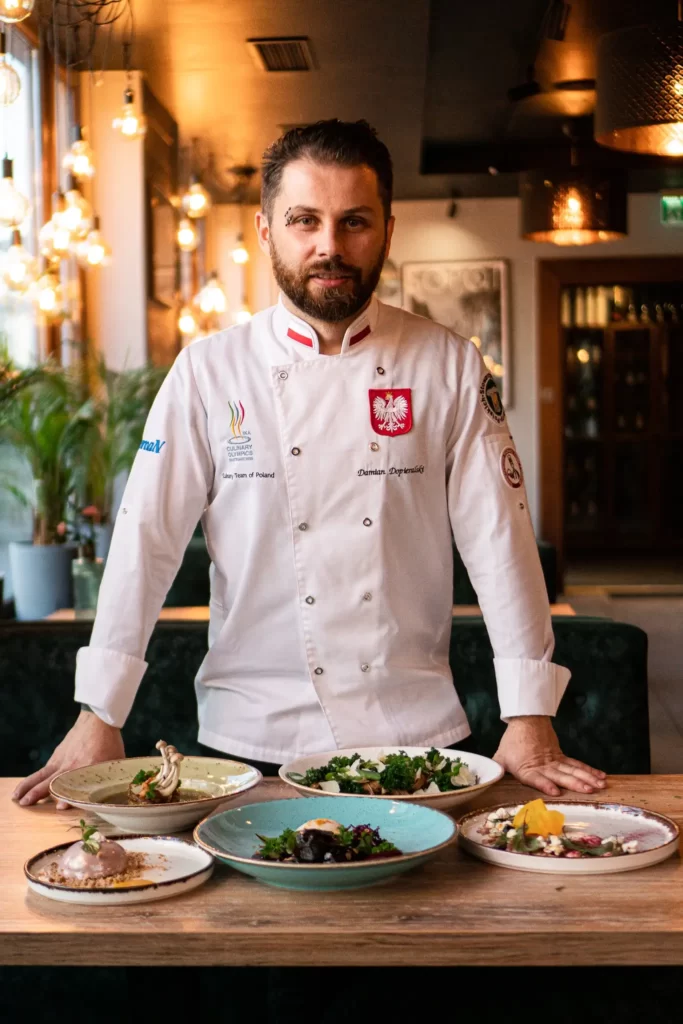Młody mężczyzna – Szef Kuchni Damian Dopieralski, stoi z rękami złożonymi na piersi nad stołem. Na stole znajduje się pięć talerzy z pięknymi daniami, przygotowanymi przez Szefa. Szef lekko się uśmiecha. W tle widać wnętrze restauracji.